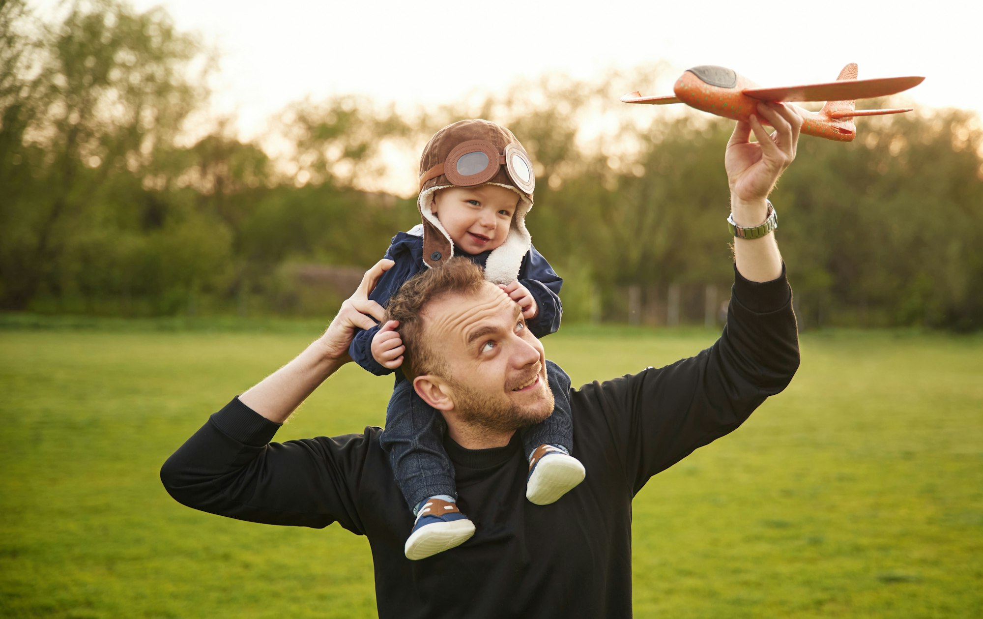 Boy is sitting on man's shoulders. Father with his little son playing with toy plane on the field