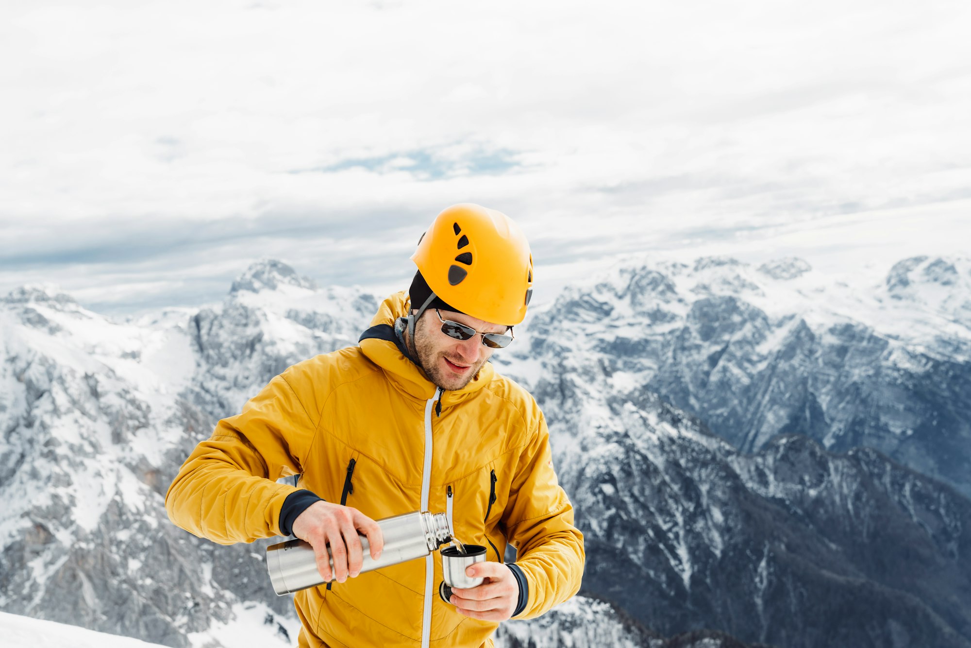 Mountaineer in a yellow jacket and helmet pouring himself some tea, view of snowy Alps in the