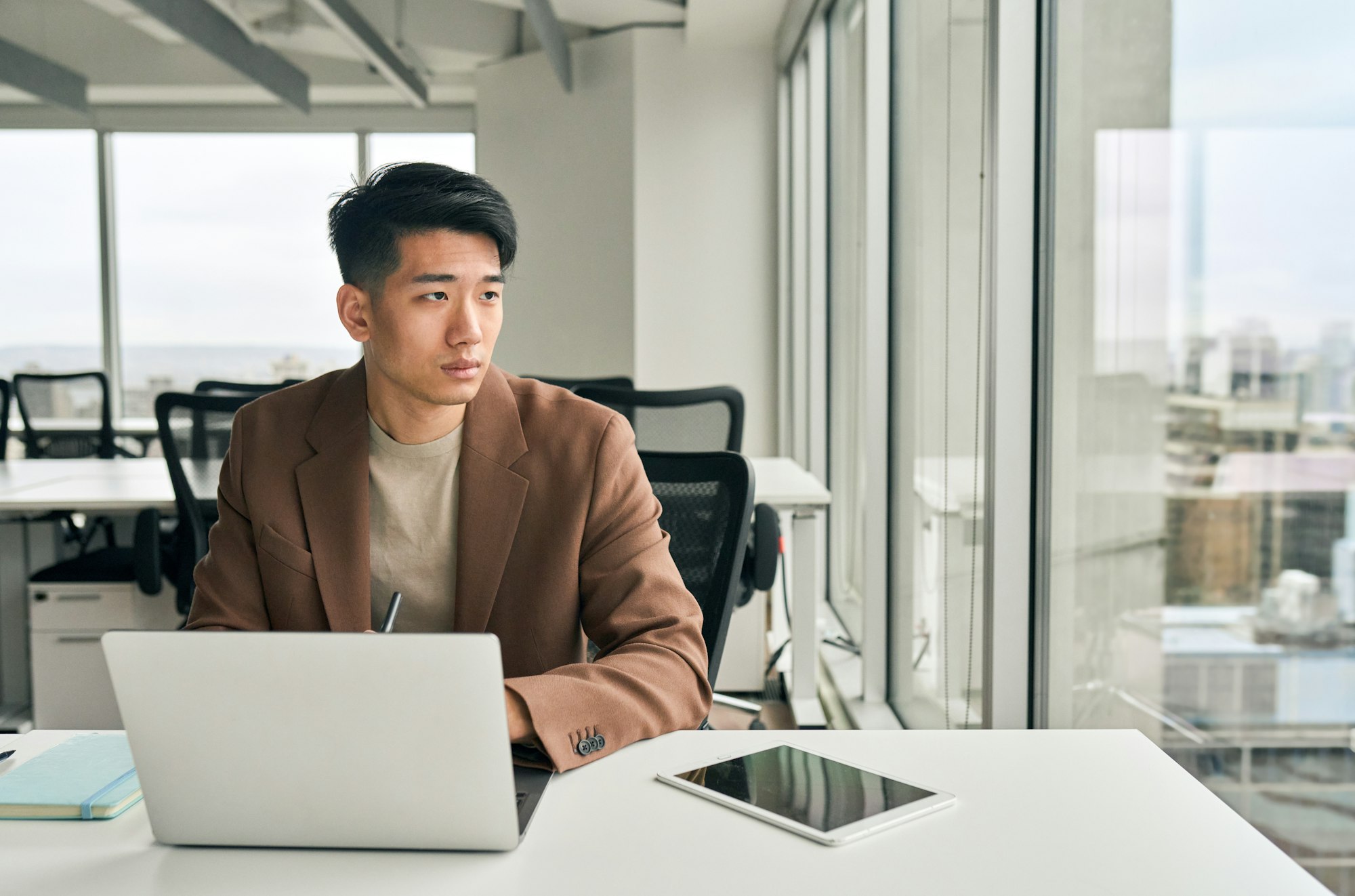 Young Asian businessman looking away thinking working on laptop in office.